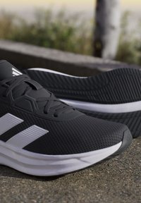 Chaussures de sport noires avec des bandes blanches et des semelles blanches épaisses reposant sur une surface en béton à l'extérieur.