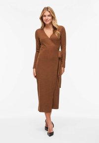 Robe portefeuille marron en tissu tricoté doux, avec manches longues, décolleté en V profond et détail de lien sur le côté. Longueur mi-mollet. Talons noirs.