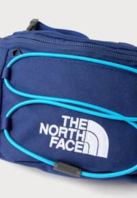 Tmavě modrá ledvinka z texturované tkaniny s jasně modrými elastickými prvky a bílým vyšívaným logem, navržená pro venkovní aktivity.