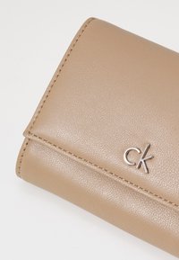 Bežová textúrovaná kožená peňaženka so strieborným logom "ck", so viditeľným prešívaním a obdĺžnikovým tvarom.
