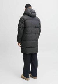 Abrigo de plumas largo y negro con capucha, secciones acolchadas, mangas anchas y un dobladillo ligeramente redondeado. Combinado con pantalones oscuros y calzado claro.