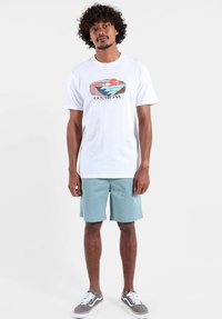 T-shirt en coton blanc avec un motif géométrique coloré et un logo sur le devant, accompagné d'un short bleu clair et de baskets grises.