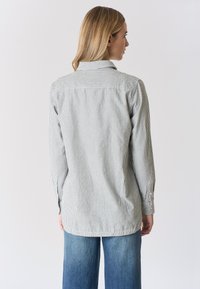 Camicia a righe grigia e bianca con bottoni, maniche lunghe e spalla posteriore; abbinata a jeans blu a gamba larga.