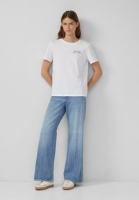 T-shirt en coton blanc avec un texte bleu sur le côté gauche de la poitrine, associé à un jean large bleu clair et des baskets beiges avec des accents blancs.