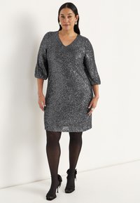 Glittrande svart klänning med V-ringning och trekvarts puffärmar, tillverkad av ett strukturerat material. Matchad med svarta klackar och strumpbyxor.