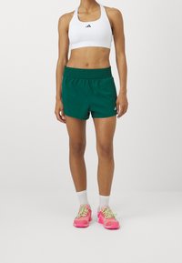Pantalones cortos deportivos verdes con cinturilla elástica, combinados con un sujetador deportivo blanco y zapatillas de colores. Calcetines cortos blancos visibles.