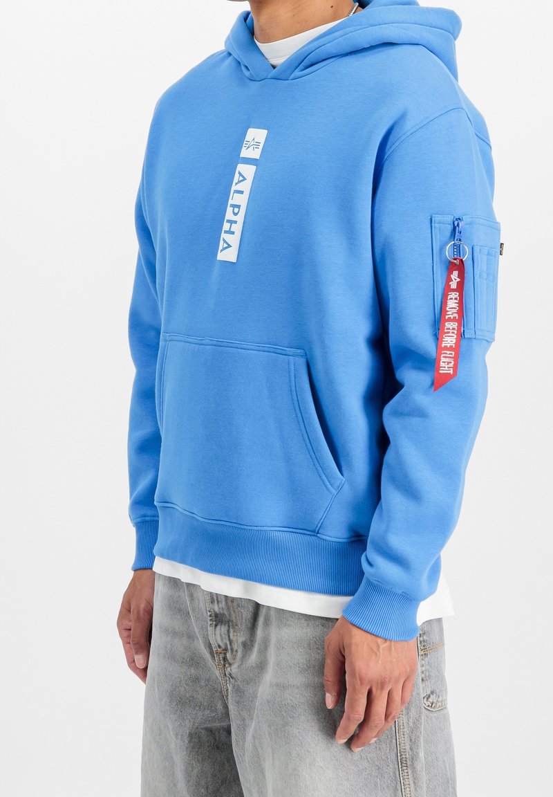 Homme portant un sweat à capuche Alpha bleu avec poche frontale et étiquette rouge "Remove Before Flight" sur la poche de la manche gauche, assorti à un jean gris clair.