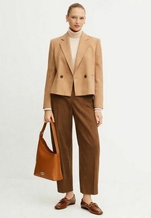 Giacca doppiopetto color cammello sopra un dolcevita crema, abbinata a pantaloni marroni su misura. Borsa e scarpe in pelle marrone con dettagli decorativi.