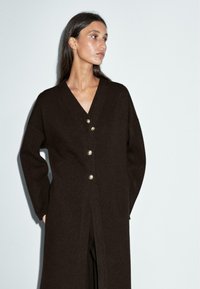 Cardigan long marron foncé en tissu doux, avec un col en V, des manches longues, et quatre boutons brillants sur le devant.