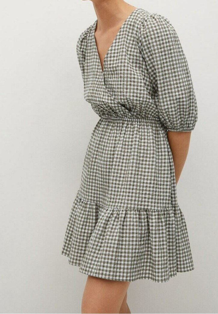 Femme portant une robe à carreaux vichy verte et blanche avec des manches bouffantes, un décolleté en V, une taille cintrée et un ourlet à volants, debout devant un fond uni.