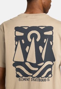Beige Baumwoll-T-Shirt mit einem großen schwarzen Grafikdesign aus dreieckigen Formen und wellenförmigen Linien, versehen mit der Marke "ELEMENT SKATEBOARD CO." am unteren Rand.
