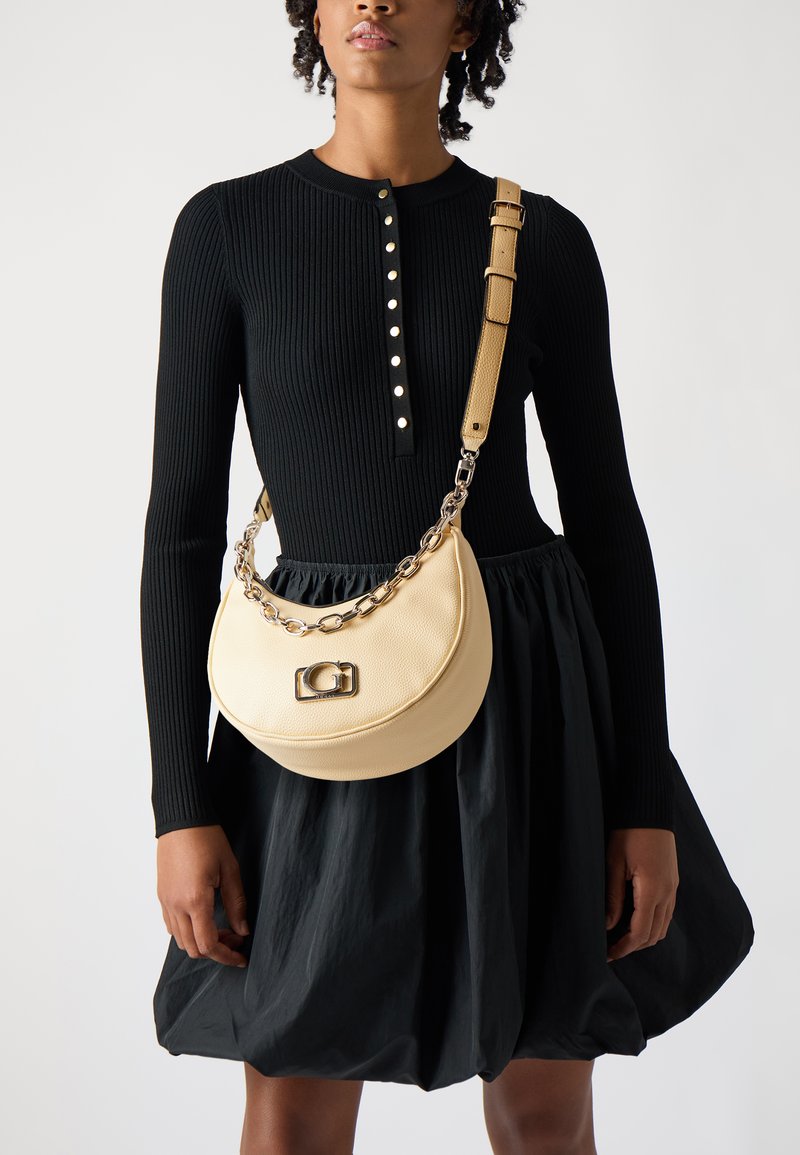 Sac à main en cuir crème de forme demi-lune, avec une chaîne en argent et un fermoir rectangulaire ; porté sur un haut à manches longues côtelé noir et une jupe.