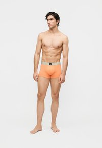 Muskelöser Mann steht barfuß in orangefarbenen Boxershorts mit grauem Bund vor hellem, schlichten Hintergrund.