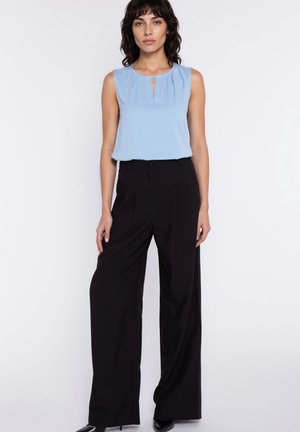 Kobieta w niebieskiej, bezrękawnikowej bluzce oraz czarnych, szerokich spodniach z wysokim stanem, stojąca na białym tle.
