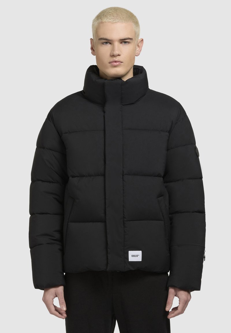 Schwarze Steppjacke mit hohem Kragen, gestepptem Design und Seitentaschen. Verfügt über ein Logo-Patch am Saum und einen Reißverschluss vorne.