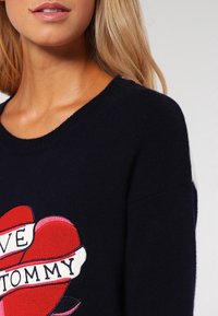 Marineblauer Strickpulli mit Rundhalsausschnitt, versehen mit einem gestickten roten Herzen und einem Banner, auf dem "LOVE TOMMY" steht. Weicher Griff, lockere Passform.