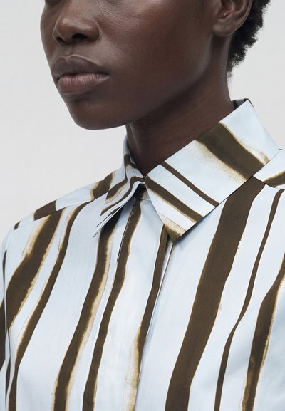 Κοντινή λήψη ενός ατόμου που φοράει πουκάμισο με γιακά με κατακόρυφες καφέ και λευκές ρίγες, εμφανίζοντας το κάτω μέρος του προσώπου και την περιοχή του λαιμού.