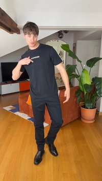 Νέος άνδρας με μαύρα ρούχα δείχνει το λογότυπο στη μπλούζα του, στέκεται σε ξύλινο πάτωμα σε μοντέρνο σαλόνι με φυτά και έπιπλα.