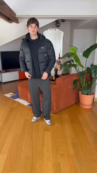 Geacă puf negre cu fermoar, purtată peste un hanorac negru. Pantaloni de trening gri și adidași argentii. Setare interioară cu o plantă și mobilier.