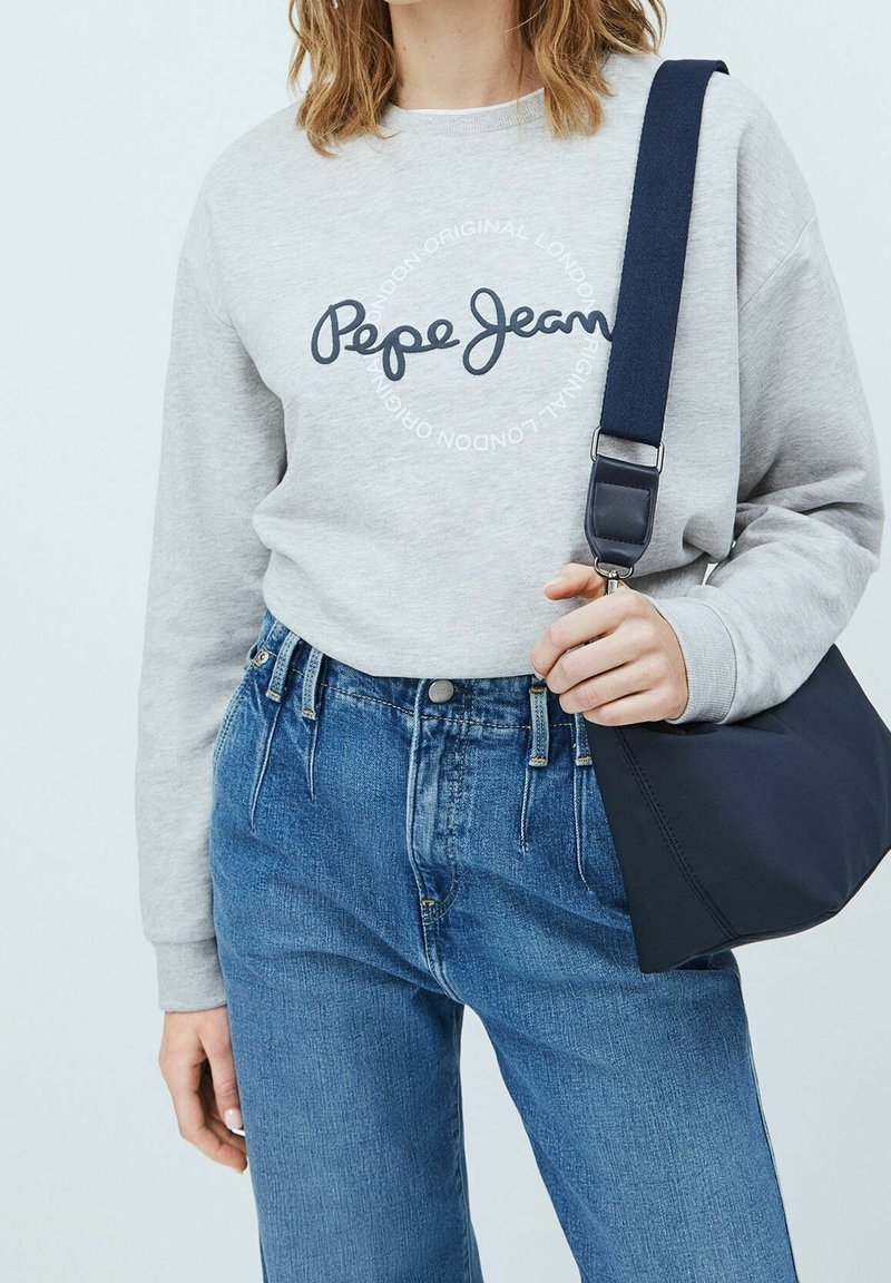 Felpa grigia con logo "Pepe Jeans", abbinata a jeans blu a vita alta. Nella mano si tiene una borsa scura con tracolla.