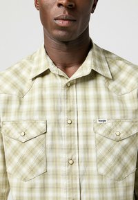 Camicia a quadri verde chiaro con bottoni, due tasche sul petto, dettagli bianchi e logo Wrangler. Presenta un colletto appuntito e bottoni a pressione.
