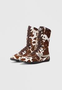 Braun-weiß gemusterte Stiefel mit Kuhflecken, Schnürung vorne, Mesh-Zunge und kontrastierenden Nähten. Schlankes Design mit strukturierter Oberfläche.