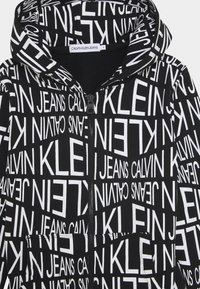 Felpa nera con zip caratterizzata da un audace testo bianco "CALVIN KLEIN JEANS" in varie oriеntazioni, con un tessuto testurizzato e tasche frontali.