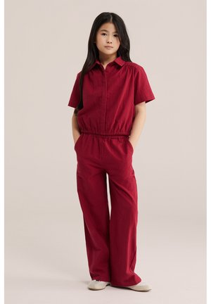 Junges Mädchen steht mit den Händen in den Taschen, trägt einen roten Kurzarm-Einteiler und weiße Schuhe vor einem einfarbigen Hintergrund.