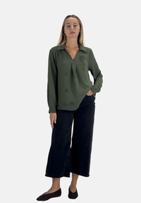 Olijfgroen blouse met lange mouwen, kraag en geplooide voorkant. Gepaard met zwarte broek met wijde pijpen en donkere platte schoenen. Eenvoudig, textured ontwerp.