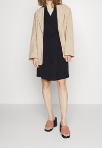 Beige oversized blazer přes černé šaty se širokým páskem, doplněné růžovými tkanými pantoflemi na blokovém podpatku. Hladká tkanina se subtilní texturou.