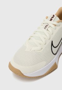 Kremaste mrežaste atletske tenisice s teksturiranom površinom, crni Swoosh logo, svijetlosive naglaske i gumu podlogu. Čipke su bijele.