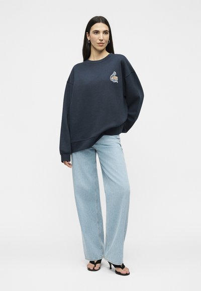 Femme debout, vêtue d'un sweat-shirt bleu marine oversize avec un petit logo sur la poitrine, un jean large bleu clair et des talons ouverts noirs.