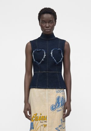 Naine kannab varrukateta tumedat teksariietusega sarnast topi, millel on narmendatud südamekujundid, ja beeži seelikut, millel on sinine ja kollane graafiline tekst ja pildid.