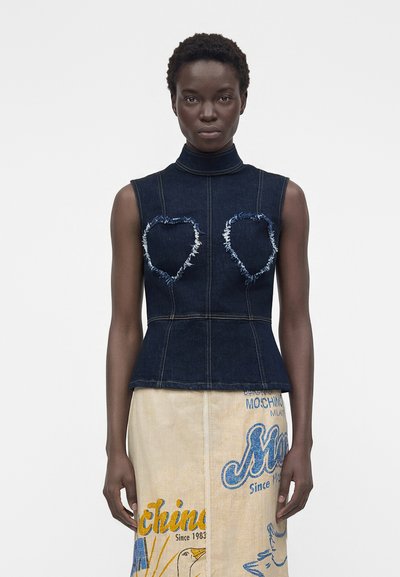Femme portant un haut sans manches en denim foncé avec des formes de cœur effilochées et une jupe beige avec des textes graphiques bleus et jaunes ainsi que des images.