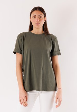 Donna con capelli lisci e scuri che indossa una t-shirt verde oliva a maniche corte e pantaloni bianchi, in piedi contro uno sfondo chiaro e uniforme.