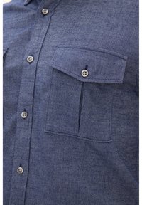 Camicia in denim blu con una trama diagonale texturizzata. Caratterizzata da una chiusura con bottoni frontale, due tasche sul petto e bottoni grigi.