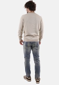 Uomo con capelli ricci che indossa un maglione beige, jeans blu sbiaditi con macchie di vernice e sneakers bianche e marroni Nike, in piedi di spalle.