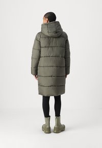 Cappotto imbottito verde oliva con cappuccio, caratterizzato da un design trapuntato orizzontale e una lunghezza fino al ginocchio. Abbinato a leggings neri e stivaletti verdi.