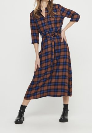 Woman wearing a brown and blue plaid midi dress with three-quarter sleeves, tied waist belt, and black ankle boots, standing with one hand on hip.