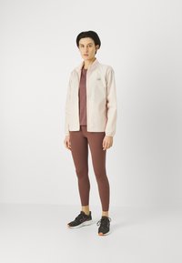 Giacca a vento rosa chiaro con polsini elasticizzati, maglietta fitted bordeaux, leggings marroni e sneaker nere con accenti arancioni.