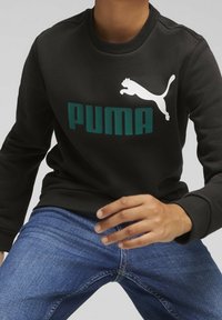 Osoba ubrana w czarną bluzę Puma z logo i niebieskie dżinsy, siedząca z uniesioną lekko do przodu jedną ręką.
