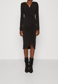 Czarna sukienka z długim rękawem i dekoltem w kształcie litery V, z marszczeniami po bokach oraz wysokim rozcięciem. Noszona z błyszczącymi czarnymi botkami za kostkę.