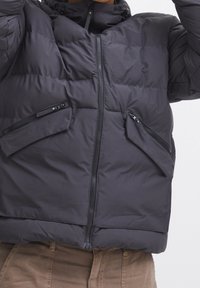 Chaqueta acolchada gris con cuello alto, cierre frontal con cremallera y dos bolsillos inclinados con cremallera. Presenta textura acolchada y tela duradera.