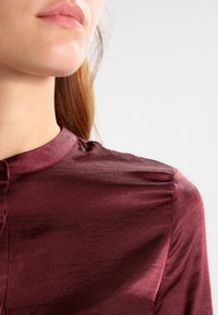 Burgundy sidenblus med rund halsringning och subtil lyster, med knäppning och lätt rynkning vid axlarna.