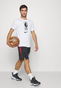 Fehér póló NBA logóval, fekete rövidnadrág piros oldalsó részletekkel, kosárlabdát tartva, fekete és fehér sportcipőt viselve, semleges háttér előtt áll.