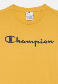 Žluté bavlněné tričko s kulatým výstřihem a tmavě modrým logem "Champion". Popis péče viditelný uvnitř límce.