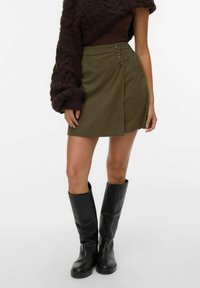 Kvinde iført en mørkegrøn omslagsnederdel med tre knapper, en mørkebrun oversized sweater og sorte knælange læderstøvler.