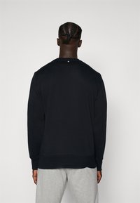 Μαύρη φούτερ με μακριά μανίκια, ριμπ μανσέτες και στρίφωμα, λεία υφή, με χαρακτηριστικό ένα μικρό ετικέτα στη πίσω πλευρά του λαιμού. Ορατές γκρίζες παντελόνες.