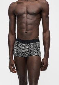 Homme torse nu portant un boxer à motifs noir et blanc avec une ceinture noire, debout devant un fond clair uni.