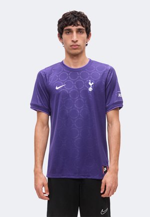 Jeune homme portant un maillot de football violet Tottenham Hotspur avec le logo Nike, se tenant devant un fond clair uni.